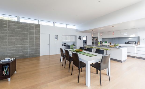 Bright, open-plan contemporary kitchen and dining area with a large white dining table, dark dining chairs, white island and cabinetry, grey tiled feature wall, pale timber flooring, and extensive glazed clerestory windows. The kitchen includes a long island with integrated seating and pendant lighting.
