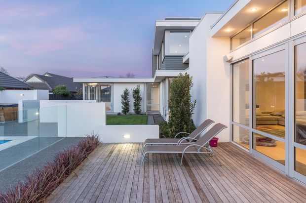 Contemporary multi-level residence viewed from a spacious timber deck at dusk. The scene features full-height glazed doors and windows, clean white rendered walls, a glass balustrade, integrated exterior lighting, landscaped planting, and a partially visible pool or water feature. Two outdoor loungers sit on the deck, while a warmly lit interior is visible through the glazing.
