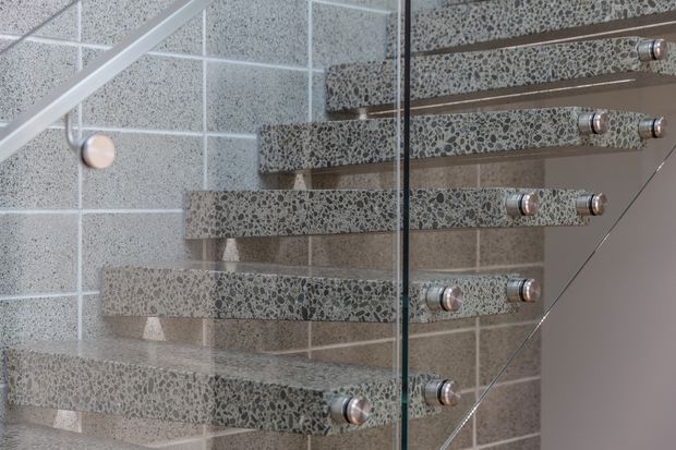 Close-up architectural view of a floating staircase with thick terrazzo-like treads, exposed round metal fixings, and a clear glass balustrade. The stair structure is set against a large-format tiled wall in a restrained neutral palette.