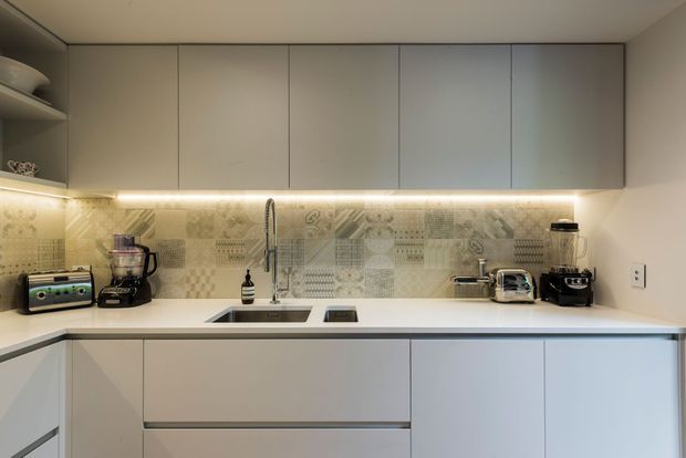 A compact modern kitchen with streamlined matte light-grey cabinetry, a white benchtop, under-cabinet lighting, and a decorative patterned tile splashback. The U-shaped work area includes an undermount sink, induction cooktop, toaster, blender, kettle, and coffee machine.