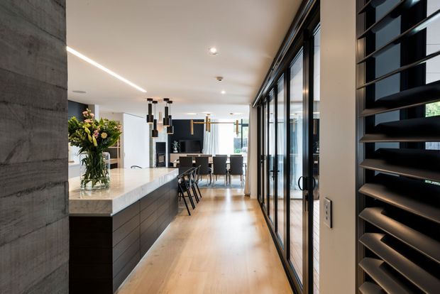 Contemporary open-plan kitchen and dining area viewed along a light timber floor. The kitchen features a large pale stone island with dark wood cabinetry, black pendant lights, and a concrete-look feature wall. Full-height black-framed glazing and a dark louvred screen line the right side, while a dining table and chairs sit beyond the island.