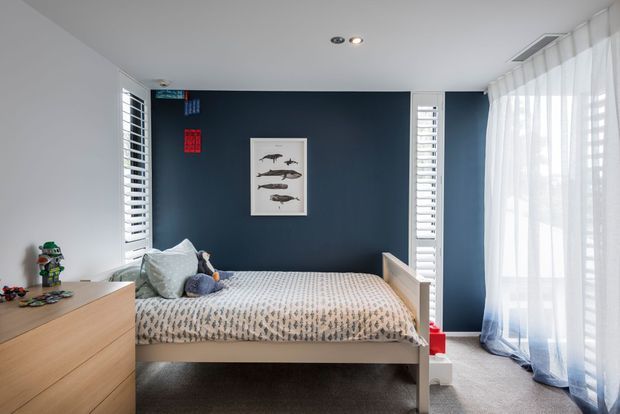 Bright, compact bedroom with a bed centered against a deep navy feature wall. White plantation shutters flank the bed, while sheer floor-to-ceiling curtains cover a large glazed opening on the right. The room includes a light timber dresser, soft grey bedding, carpeted flooring, recessed ceiling lights, and playful toy accents.