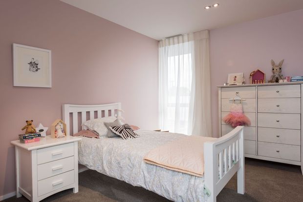 A soft, pastel children's bedroom with a white spindle-style bed, white bedside table, tall white dresser, and layered bedding. Pale pink walls, sheer floor-length curtains, recessed ceiling lights, plush toys, and small decorative accessories create a warm and playful atmosphere.