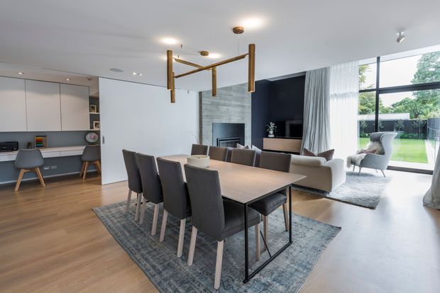A bright, open-plan contemporary living and dining area with a modern kitchen at left. The room features warm timber flooring, a large dining table with upholstered chairs, a neutral sofa seating area, a dark fireplace wall with a television, full-height glazing, and a sculptural brass-toned pendant light.