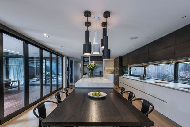 A contemporary open-plan kitchen and dining area with a long dark timber dining table, black dining chairs, a pale stone kitchen island, white lower cabinetry, and dark timber wall cabinetry. Multiple black pendant lights hang above the island, while extensive floor-to-ceiling glazing opens to a deck or outdoor area. The space has clean lines, integrated appliances, and a refined neutral palette.