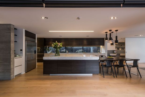 Open-plan contemporary kitchen and dining area with a large dark timber island, pale stone benchtop, integrated dark cabinetry, stainless-steel appliances, timber flooring, and a black dining table with pendant lights. A textured concrete or stone column frames the left side, while a long horizontal window provides natural light behind the kitchen.