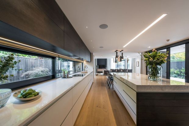 A spacious contemporary kitchen with a long central island, extensive white cabinetry and pale stone benchtops. Dark timber-toned overhead cabinetry contrasts with the light finishes, while integrated linear lighting, pendant lights, and large glazed windows create a bright, refined interior. A dining and living area is visible beyond the kitchen, with landscaped greenery seen through the windows.
