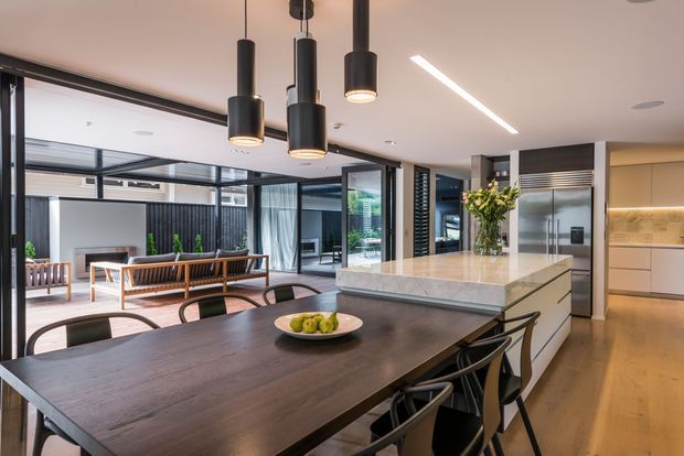 Contemporary open-plan kitchen and dining space with a large dark timber dining table, upholstered dining chairs, a central island with a pale stone benchtop, integrated cabinetry, and three black pendant lights. Full-height black-framed glazing connects the interior to a courtyard or outdoor living area, while warm timber flooring and soft neutral finishes create a refined, inviting atmosphere.