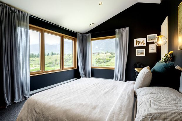 A stylish bedroom with dark navy-black walls, a large upholstered bed dressed in light textured bedding, and expansive timber-framed corner windows overlooking a green landscape. Full-height grey curtains, framed wall art, and a spherical pendant light create a refined, contemporary atmosphere.