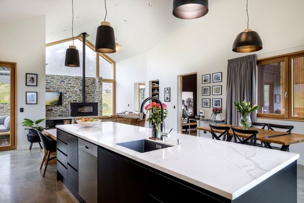 Open-plan contemporary kitchen and dining area with a large white stone island, dark cabinetry, integrated sink and cooktop, pendant lighting, timber-framed windows, and a dining table. A stacked-stone feature wall and living area are visible beyond the kitchen.