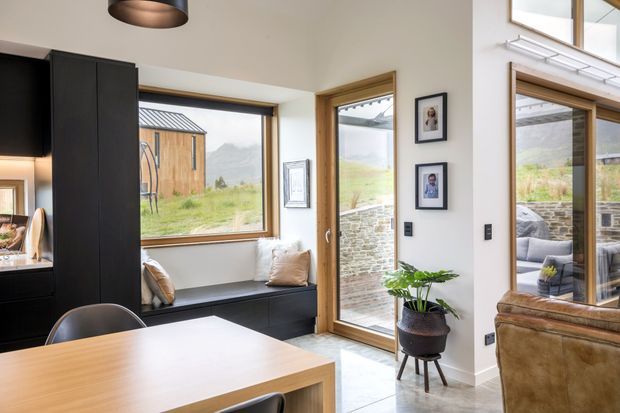 A bright, modern open-plan dining and living area with a timber dining table, black built-in cabinetry and window seat, large timber-framed windows, and a glazed timber-framed door opening to an outdoor terrace. Natural stone paving, framed photographs, indoor planting, and views toward a grassy rural landscape and hills are visible.