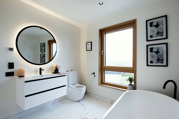 Bright contemporary bathroom featuring a floating white vanity with black horizontal detailing, black tapware, a round black-framed mirror, wall-hung toilet, freestanding white bathtub, marble-look tiled floor, timber-framed window, and monochrome artwork.