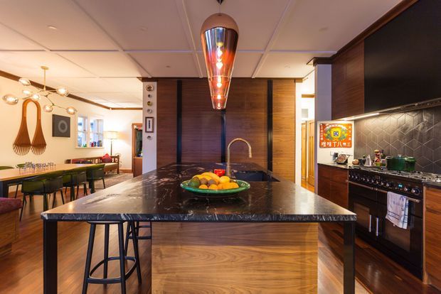 Warm, richly detailed open-plan kitchen viewed from the island. The room features dark timber cabinetry and wall panelling, a large dark stone island benchtop with integrated seating, a brass-toned tap, patterned dark tile splashback, and a professional freestanding range. A dining area and living space extend beyond the kitchen, with pendant and track lighting creating an inviting atmosphere.