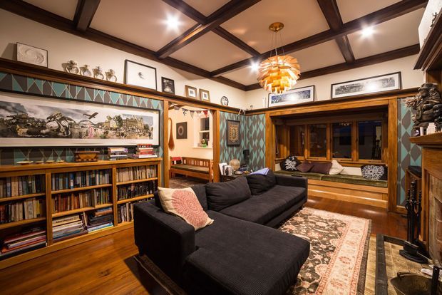 Warm, richly detailed traditional living room with dark timber ceiling beams, extensive built-in bookshelves, wood flooring, patterned wall treatments, framed artwork, and a large black sectional sofa arranged around a rug. A deep timber-framed window seat and decorative fireplace surround contribute to the cosy, collected character, while a sculptural pendant provides a focal point.