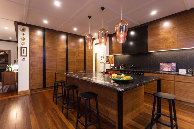 A sophisticated contemporary kitchen with extensive dark timber cabinetry and wall panelling, a large black marble or stone island with seating for several people, a dark hexagonal tile splashback, integrated appliances, and a row of decorative copper pendant lights. Warm timber flooring and recessed ceiling lights create a rich, inviting atmosphere. An adjoining dining or living area is visible at left.