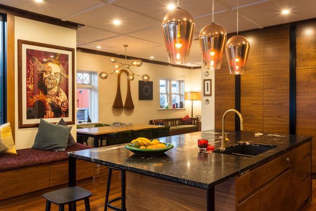 Warm, richly detailed kitchen interior centred on a large dark stone island with integrated sink and curved brass tap. The room features extensive dark timber cabinetry and wall panelling, a dining area, pendant lights with metallic shades, a built-in bench seat, framed artwork and decorative wall elements.