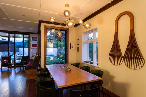 A warm, refined dining room centered on a long rectangular timber dining table with dark chairs. The room features polished timber flooring, cream walls, a decorative dark timber ceiling beam, divided-pane windows, large glazed doors opening toward an outdoor area, and a sculptural multi-globe pendant light. A distinctive paired wall sculpture adds an artistic accent.