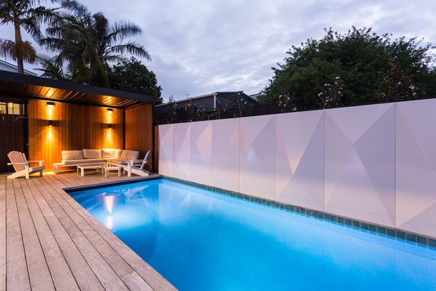 A contemporary backyard pool area at dusk, featuring a long rectangular swimming pool surrounded by a timber deck. A timber-lined covered outdoor lounge with built-in lighting and light-coloured seating sits at one end, while a geometric pale privacy fence encloses the space. Tropical palms and dense planting rise above the boundary.