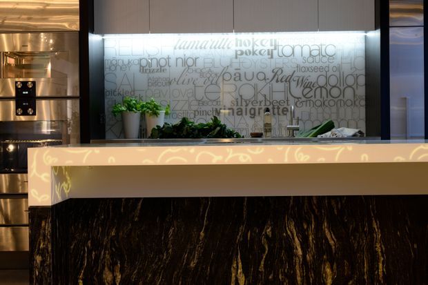 A contemporary kitchen island with a dramatic dark veined stone-clad base and a thick illuminated cream-toned benchtop. Behind it is a stainless-steel cooking wall and a backlit glass splashback featuring printed typography, with small potted plants adding greenery.