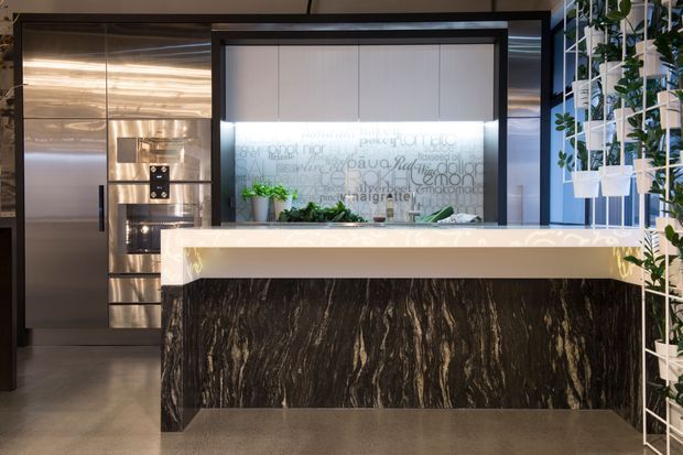 A contemporary reception or service counter dominates the image, with a dramatic dark veined stone-faced base, a pale illuminated benchtop, and integrated concealed lighting. Behind it are metallic built-in cabinets and display shelving, while a translucent graphic-glazed partition and a vertical plant trellis add visual interest. The space has a polished commercial interior fit-out with a dark, refined material palette.