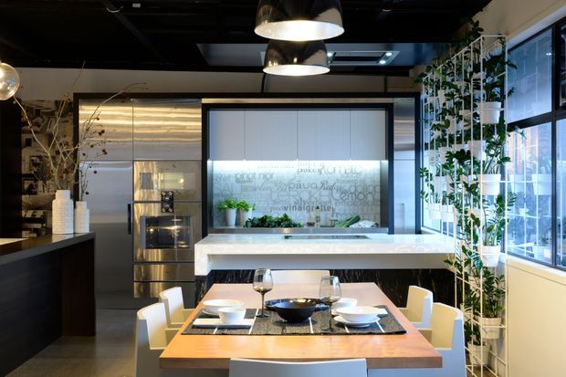 A contemporary open-plan kitchen and dining area with a large white island, integrated stainless-steel appliances, dark cabinetry, and a timber dining table set for six. Floor-to-ceiling windows line one side, complemented by a tall indoor planted screen. Black ceilings, pendant lights, and a translucent graphic splashback create a refined, high-contrast interior.
