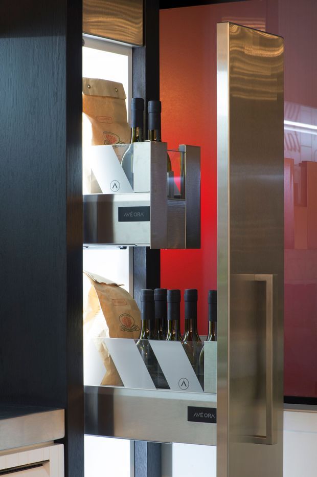 A close-up view of a refined retail display fixture showcasing wine bottles and packaged goods. The display uses brushed metal shelving and framing, dark textured wall panels, illuminated shelving niches, and a bold red backing panel, creating a premium commercial merchandising environment.
