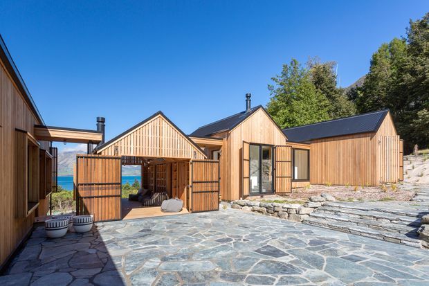 A contemporary timber-clad single-storey building complex arranged around a broad irregular stone-paved courtyard. The composition includes steep gabled volumes with dark metal roofs, vertical timber screening and gates, large glazed doors, and stepped rough-stone landscaping. Trees and a glimpse of blue water are visible beyond the buildings.