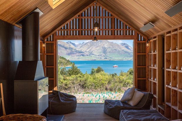 A rustic timber-lined gabled living space with a dramatic full-width opening framing a turquoise alpine lake and mountains. The room includes relaxed beanbag-style seating, a dark freestanding fireplace, built-in timber shelving, exposed slatted rafters, and timber decking, creating a warm cabin-like retreat.