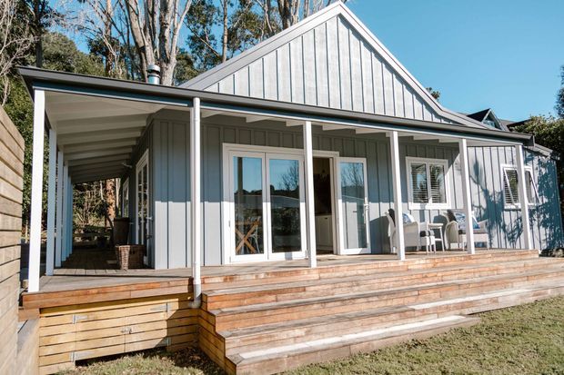 Exterior view of a single-storey home with a prominent gabled roof, grey vertical weatherboard cladding, white trim, covered verandah, and broad timber decking and steps. The house sits in a wooded suburban or bush setting.