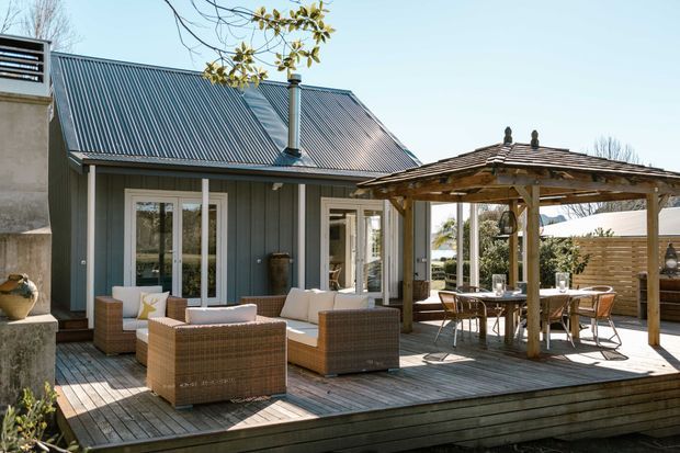 Exterior view of a coastal-style home with a grey corrugated metal gable roof, vertical grey cladding, white-framed glazed doors, and a raised timber deck. The deck includes wicker outdoor seating and a timber pergola with a shingle-like roof over an outdoor dining area.