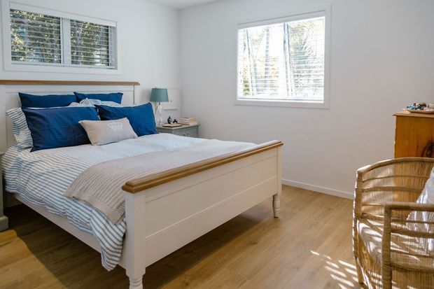 Bright, comfortable bedroom with a white paneled bed, blue and white striped bedding, timber flooring, plantation shutters, and a woven rattan chair. Natural light fills the room through two windows, creating a relaxed coastal feel.