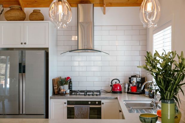 A compact, warm kitchen with white shaker-style cabinetry, a stainless-steel refrigerator and rangehood, a white subway-tile splashback, and a stainless-steel gas cooktop and sink. Timber ceiling boards and exposed timber beams add warmth, while glass pendant lights, countertop appliances, potted greenery, and ceramic vessels provide decorative detail.