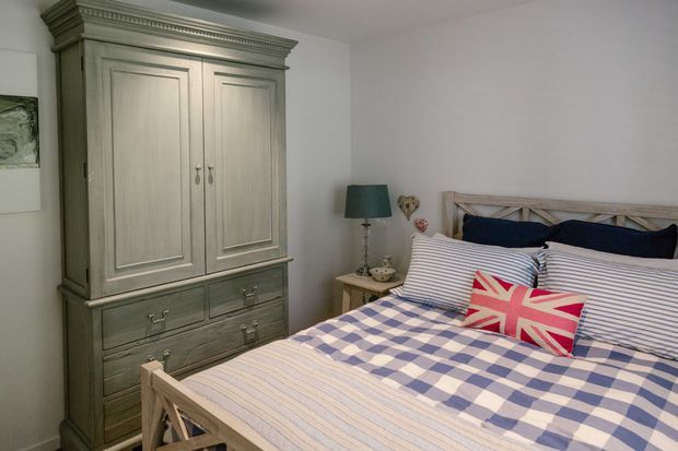 A cozy traditional bedroom with a large painted timber armoire, a timber bed featuring a cross-braced headboard, striped bedding, a blue-and-white checked throw, and a Union Jack accent cushion. Soft neutral walls and classic moulded cornice contribute to the relaxed, country-inspired character.
