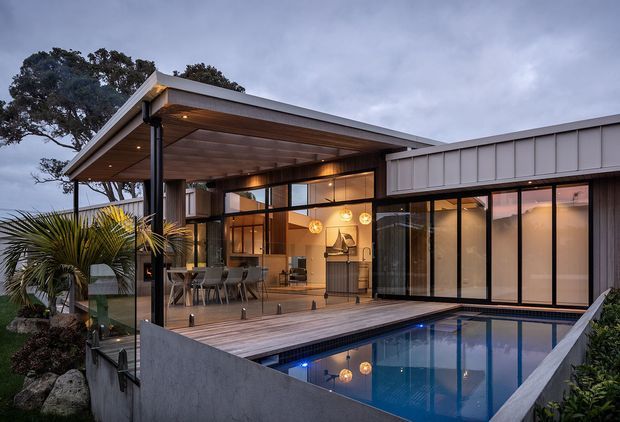 Contemporary single-storey residence viewed from the garden, featuring a broad low-pitched roof, timber-lined covered outdoor entertaining area, extensive glazing, and a rectangular swimming pool adjoining the deck. Warm interior lighting reveals an open-plan living or dining area and kitchen/bar joinery inside. The exterior uses dark vertical cladding, timber decking, concrete pool walls, and slender dark window frames, creating a refined indoor-outdoor connection at dusk.