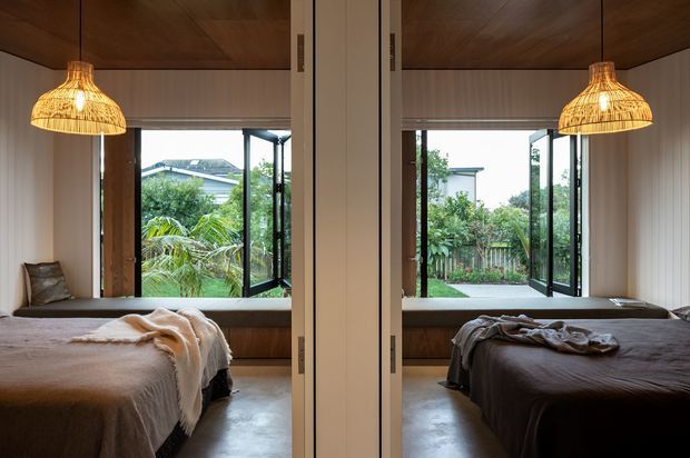A symmetrical view into two mirrored bedroom spaces, each with a bed, built-in window seat, open black-framed glazing, warm timber ceiling, and woven pendant light. The bedrooms overlook a leafy garden and neighbouring homes, with a calm, natural material palette of timber, neutral bedding, and concrete-like flooring.