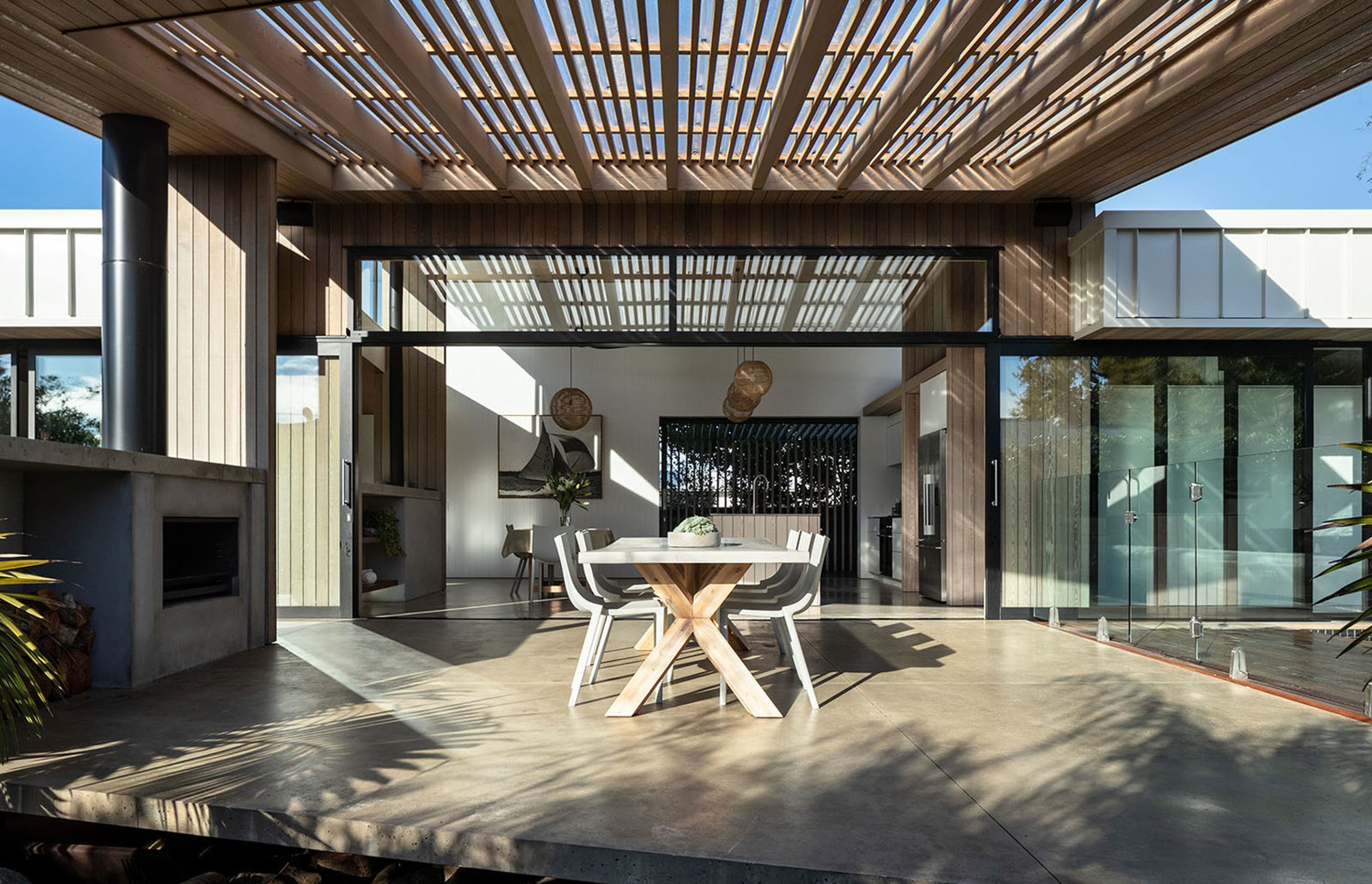 The slatted canopy creates filtered light over the outdoor room. An outdoor fireplace provides warmth on cooler evenings.