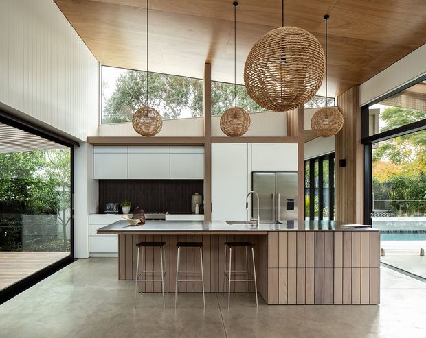 A spacious contemporary kitchen with a large island and integrated seating, set beneath a warm timber-lined ceiling. Full-height glazing connects the room to an outdoor pool area and leafy landscape. The kitchen features pale flat-panel cabinetry, a dark textured splashback, a light stone benchtop, timber island cladding, stainless-steel appliances, and woven pendant lights. Exposed timber posts and clerestory glazing emphasize the airy, indoor-outdoor character.