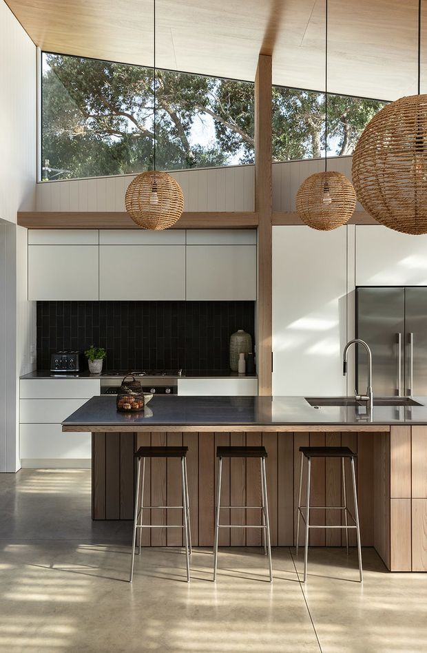 A bright, open-plan contemporary kitchen with a large island, three stools, pale integrated cabinetry, a black vertical-tile splashback, stainless-steel appliances, and a dark benchtop. Full-height glazing brings in views of trees, while exposed timber ceiling lining and a timber structural post add warmth. Woven rattan pendant lights hang above the island.