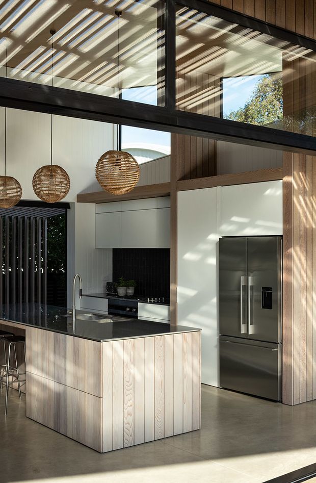 A bright, double-height contemporary kitchen with a large island clad in pale vertical timber boards and topped with a dark stone benchtop. White flat-panel cabinetry, a black tiled splashback, integrated cooking appliances, and a stainless-steel refrigerator line the rear wall. Tall black-framed glazing and clerestory windows bring in sunlight, while woven pendant lights hang above the island. The space combines warm timber finishes with polished concrete flooring and strong indoor-outdoor visual connections.