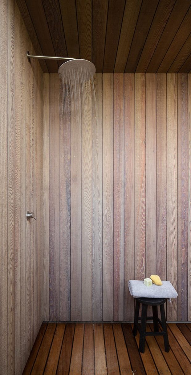 A compact walk-in shower room fully lined with vertically oriented timber boards on the walls and dark timber planks on the ceiling and floor. A large round rainfall showerhead and wall control are mounted on the left wall. A small black stool with a folded towel and soap sits in the corner. The space has a warm, rustic, natural character with minimal fittings.