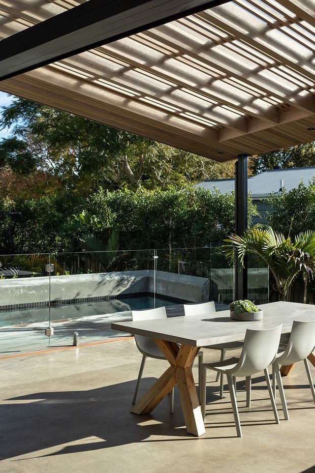 A contemporary covered outdoor dining area beside a swimming pool. The space features a large concrete dining table with timber trestle-style supports, white dining chairs, a timber-lined pergola roof with translucent panels, dark structural beams and posts, glass pool fencing, and lush garden planting surrounding the pool.