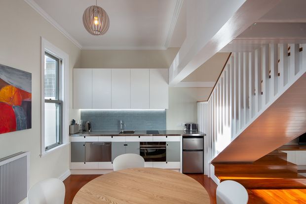 A bright, compact open-plan kitchen and dining area beneath a staircase. The kitchen features streamlined white wall cabinetry, pale blue-grey tiled splashback, dark benchtops, integrated appliances and stainless-steel fittings. A round timber dining table with white chairs occupies the foreground, while a sculptural timber stair with white vertical balustrades rises along the right side. White walls, ceiling cornices and a contemporary pendant light create a clean, warm interior.