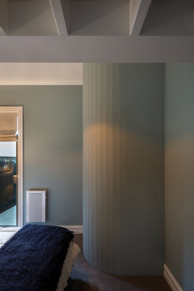A moody bedroom interior with muted blue-grey walls, white decorative cornices and exposed ceiling beams. The lower-left foreground shows part of a bed with dark navy bedding, while a tall built-in wardrobe or paneled storage wall dominates the right side. A white radiator sits beneath the wall trim, and a doorway or mirror at left provides a partial reflection of adjacent cabinetry. The room has traditional detailing paired with restrained contemporary styling and soft, warm lighting.