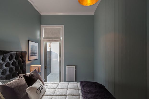 A compact, moody bedroom with deep blue-grey walls, a tall tufted upholstered headboard, and a quilted bed dressed in neutral bedding with a dark throw. White-painted trim surrounds a glazed doorway, while a slim radiator, bedside lamp, framed artwork, and full-height vertical wall panelling complete the room. Warm pendant lighting adds contrast to the cool palette.