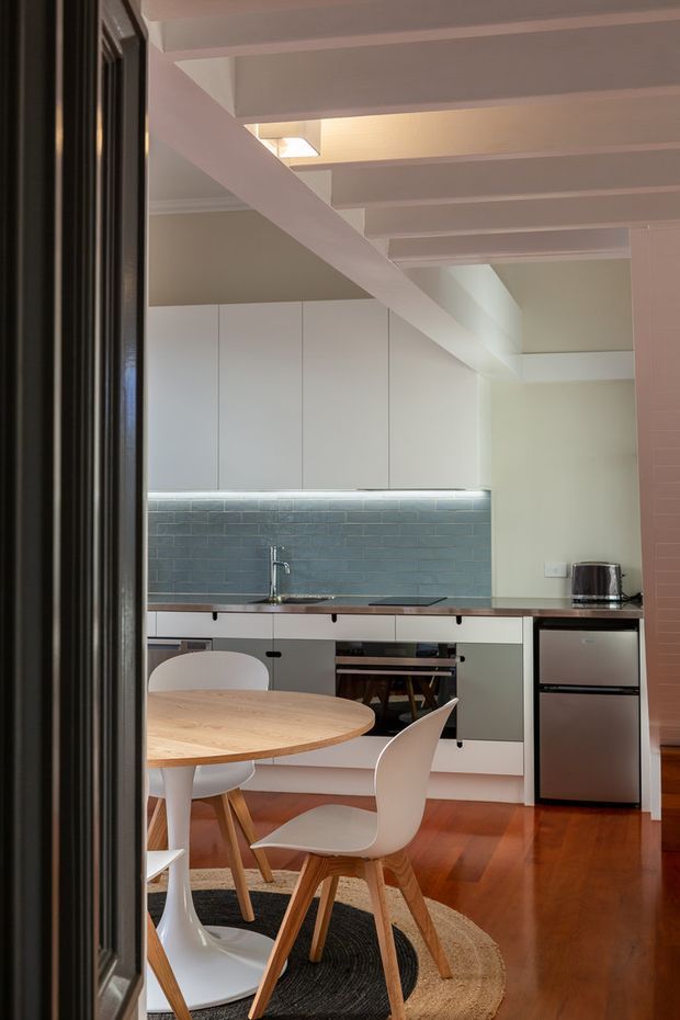 Compact open-plan kitchen and dining area viewed through a dark framed doorway. The kitchen has flat-front white and pale grey cabinetry, a blue-grey rectangular tile splashback, integrated appliances, and a slim dark benchtop. A round timber dining table with white molded chairs occupies the foreground. Warm timber flooring, exposed white ceiling beams, and concealed under-cabinet lighting create a warm contemporary interior.