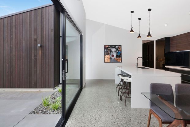 Contemporary open-plan kitchen and dining area viewed from a glazed courtyard edge. The room features a white island with integrated sink and stools, dark timber cabinetry, black-framed full-height glazing, pendant lights, polished concrete or terrazzo-like flooring, and a compact dining setting. A dark vertical timber-clad exterior volume and landscaped gravel strip are visible outside.