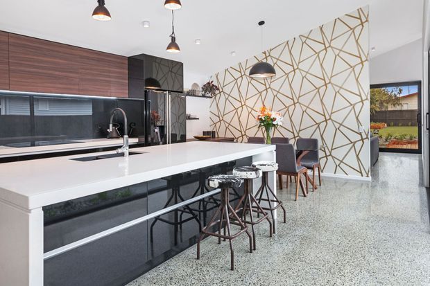 Contemporary open-plan kitchen and dining area with a large white waterfall island, glossy dark cabinetry, timber-look wall cabinetry, pendant lighting, patterned geometric feature wall, bar stools, and terrazzo-style flooring. An outdoor area is visible through the doorway.