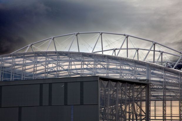 Exterior view of a large commercial or industrial structure with a dramatic curved steel truss roof and extensive translucent glazing or cladding. Dark vertically ribbed wall panels form the lower facade, while the exposed structural framing dominates the upper portion beneath an overcast sky.
