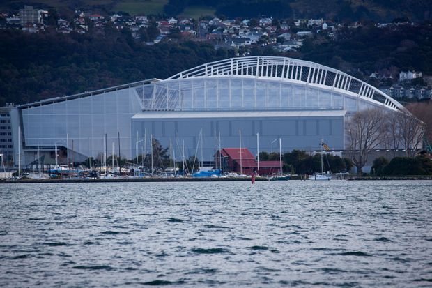 Exterior view across a body of water toward a large waterfront commercial or events building with a distinctive arched roof structure and translucent or pale cladding. Boats and marina infrastructure sit in front of the building, with a densely settled, hilly urban landscape behind it.