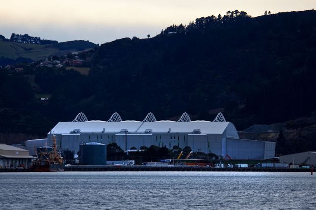 Exterior waterfront view of a large industrial or commercial building with a long low white facade, repeated arched roof structures, and a substantial cylindrical tank. The building sits at the edge of a body of water, backed by steep, densely wooded hills and scattered hillside housing. The image is photographed in subdued, overcast light.